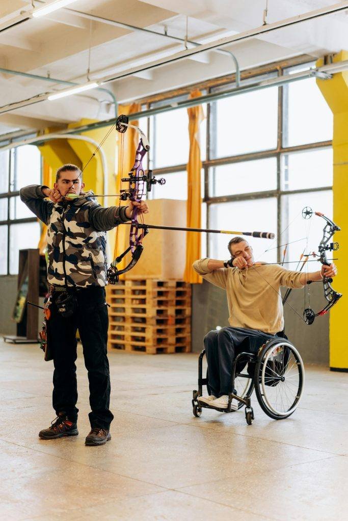 Two male archers practicing indoors, one in a wheelchair, showcasing adaptive sports' strength and focus.